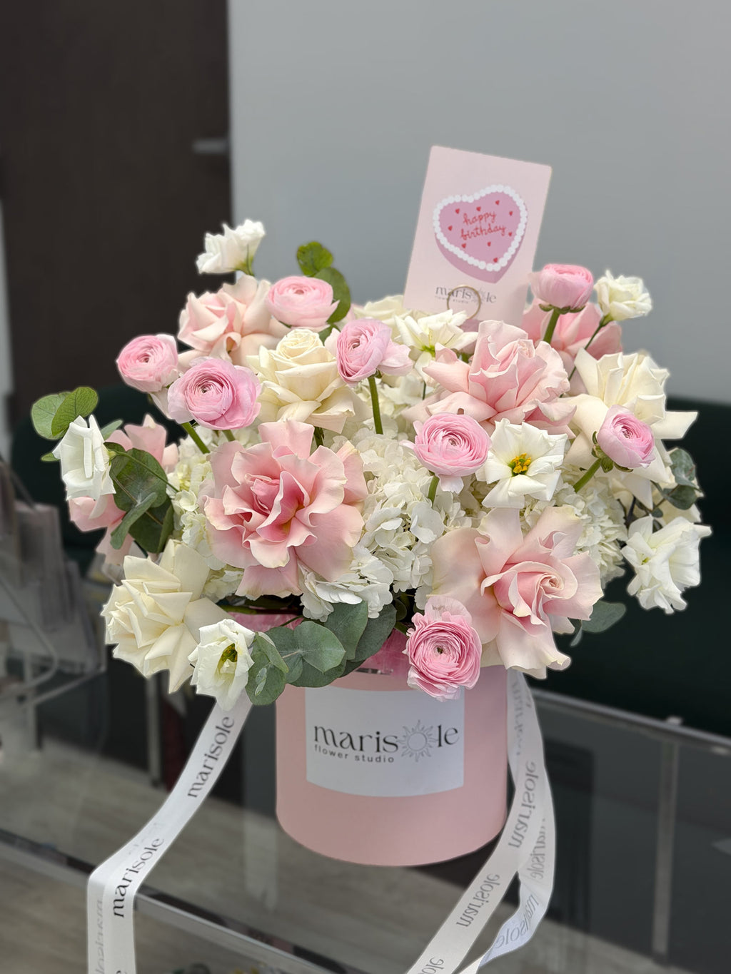 A pink round flower box filled with a bouquet of pink and white roses, white carnations, and green eucalyptus leaves, with a pink heart-shaped card and white ribbon printed with the brand name 'marisole flowers'.