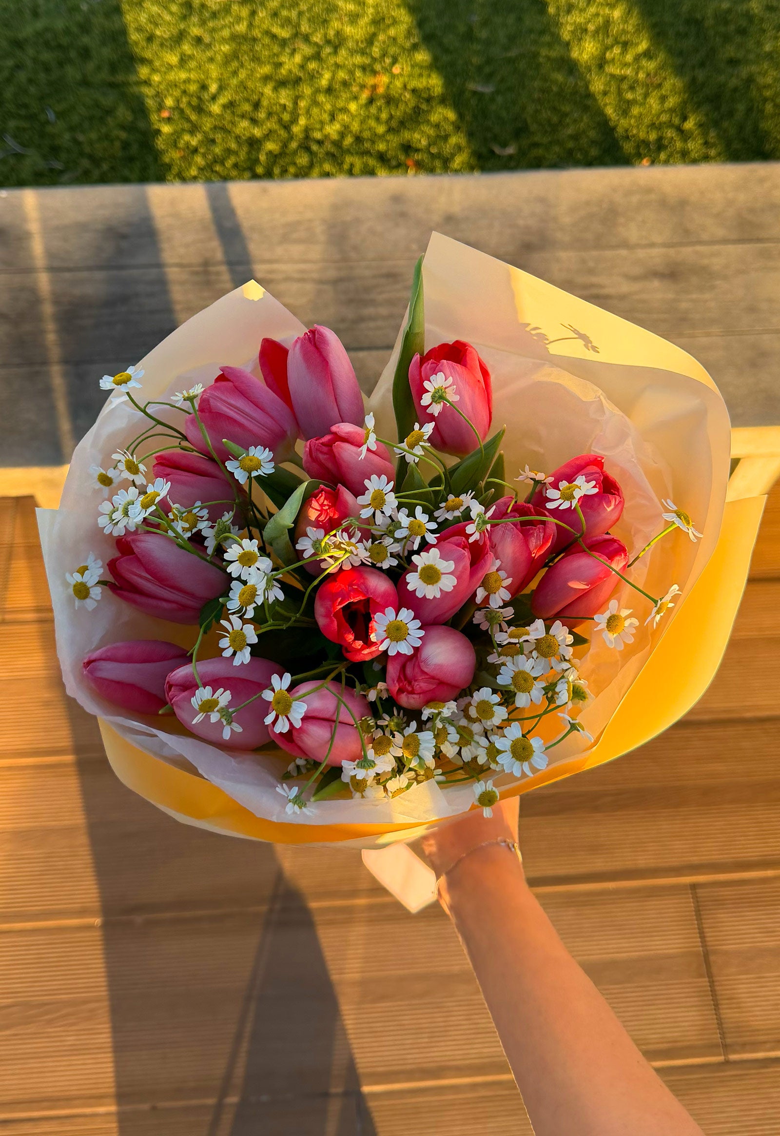 Bouquet of pink tulips and white flowers wrapped in yellow paper, held by a person outdoors.