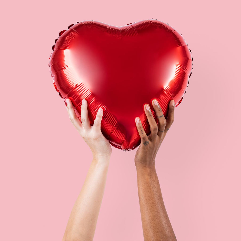 Red heart-shaped balloon held by hands against a pink background