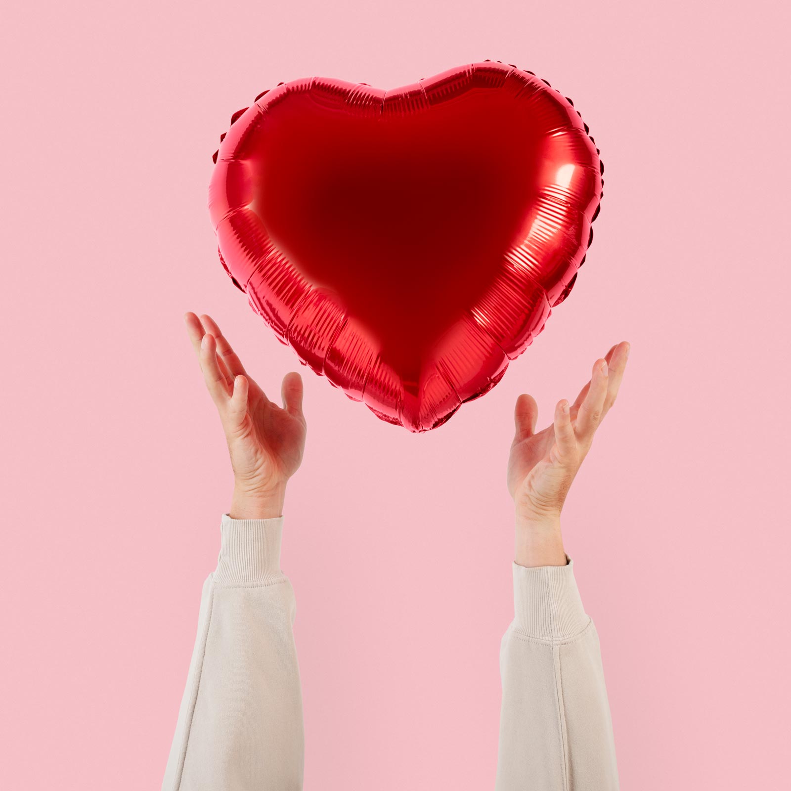 Red heart-shaped balloon held by two hands against a pink background