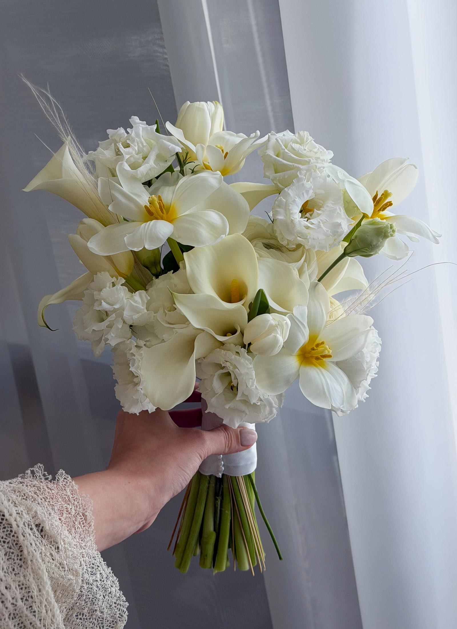 A hand holding a bridal bouquet of white calla lilies, roses, lisianthus, and white peonies with green stems, set against a soft gray curtain background.