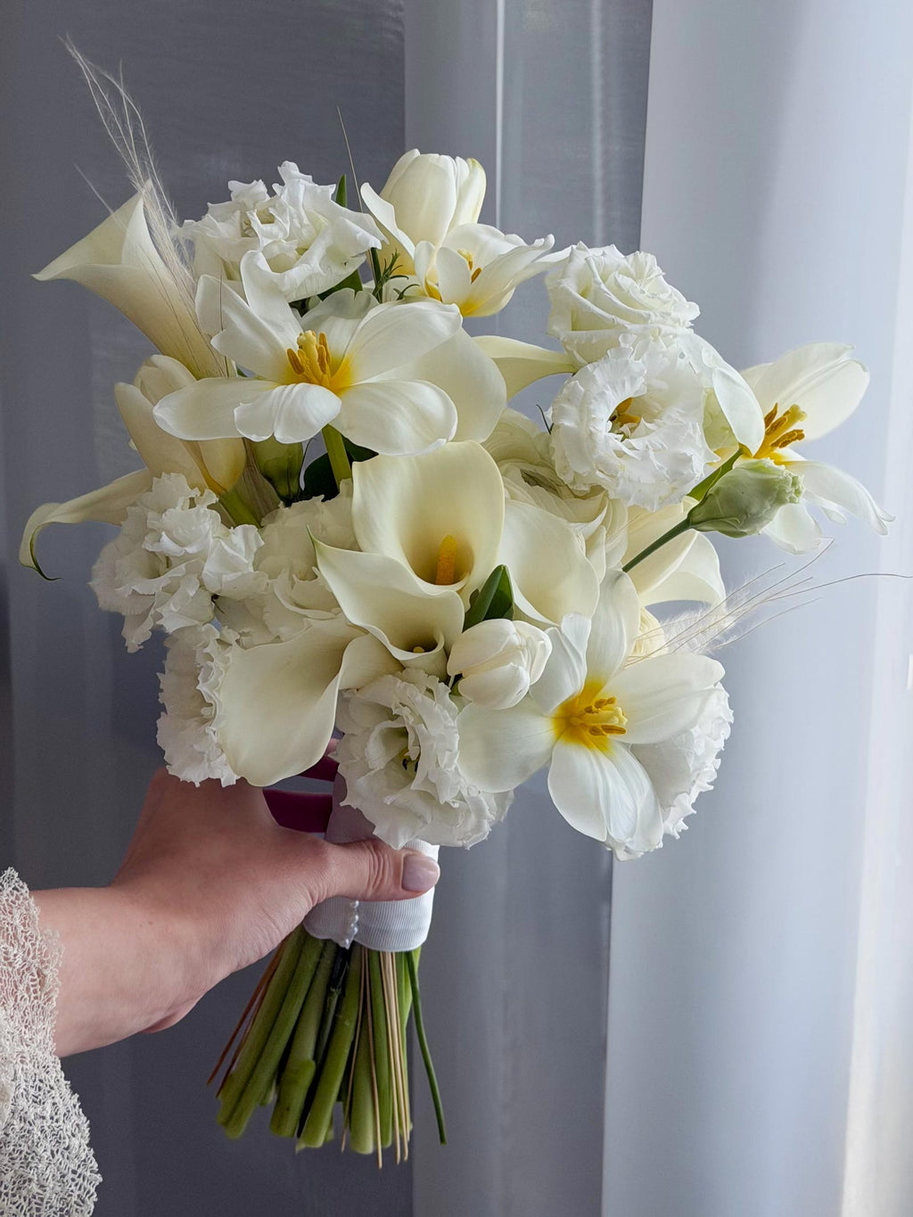 A hand holding a white floral bouquet featuring calla lilies, roses, carnations, and lisianthus with green stems, set against a soft gray curtain background.