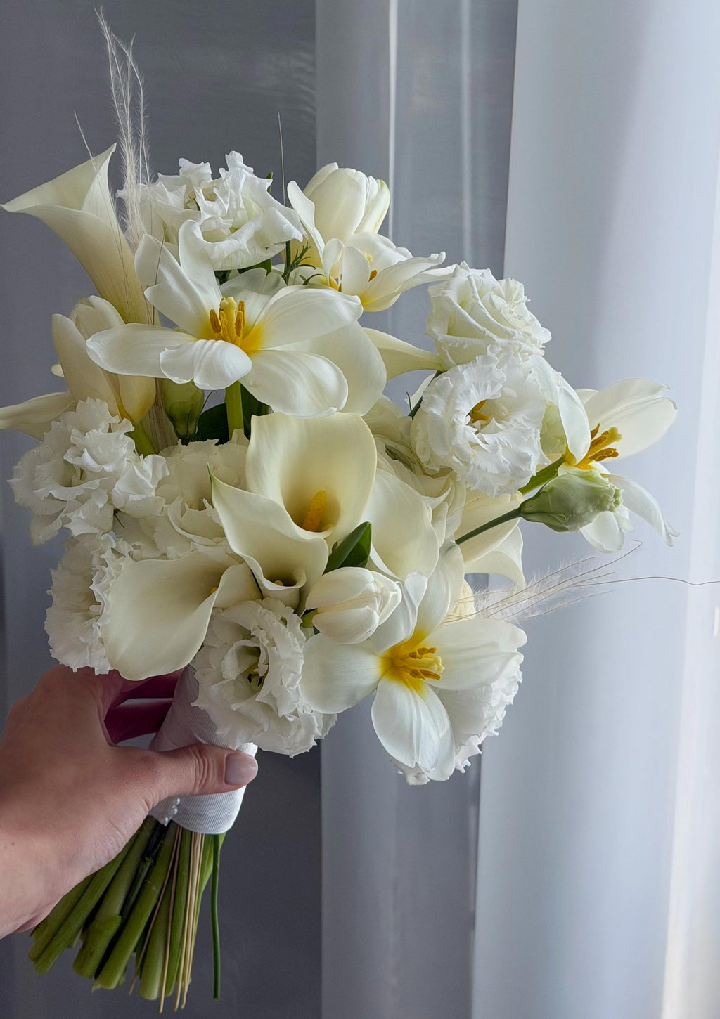 A hand holding a bouquet of white and cream flowers, including calla lilies, lisianthus, and carnations, with green stems, set against a soft gray curtain background.