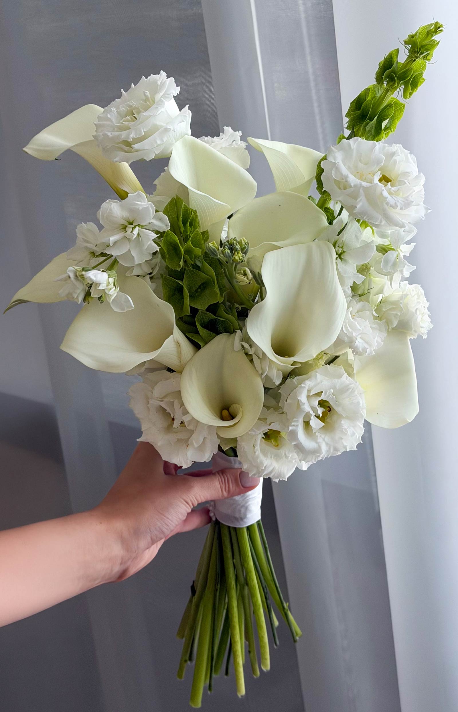 A hand holding a fresh bridal bouquet of white calla lilies, white lisianthus, and white stock flowers with green stems, set against a soft gray curtain background.