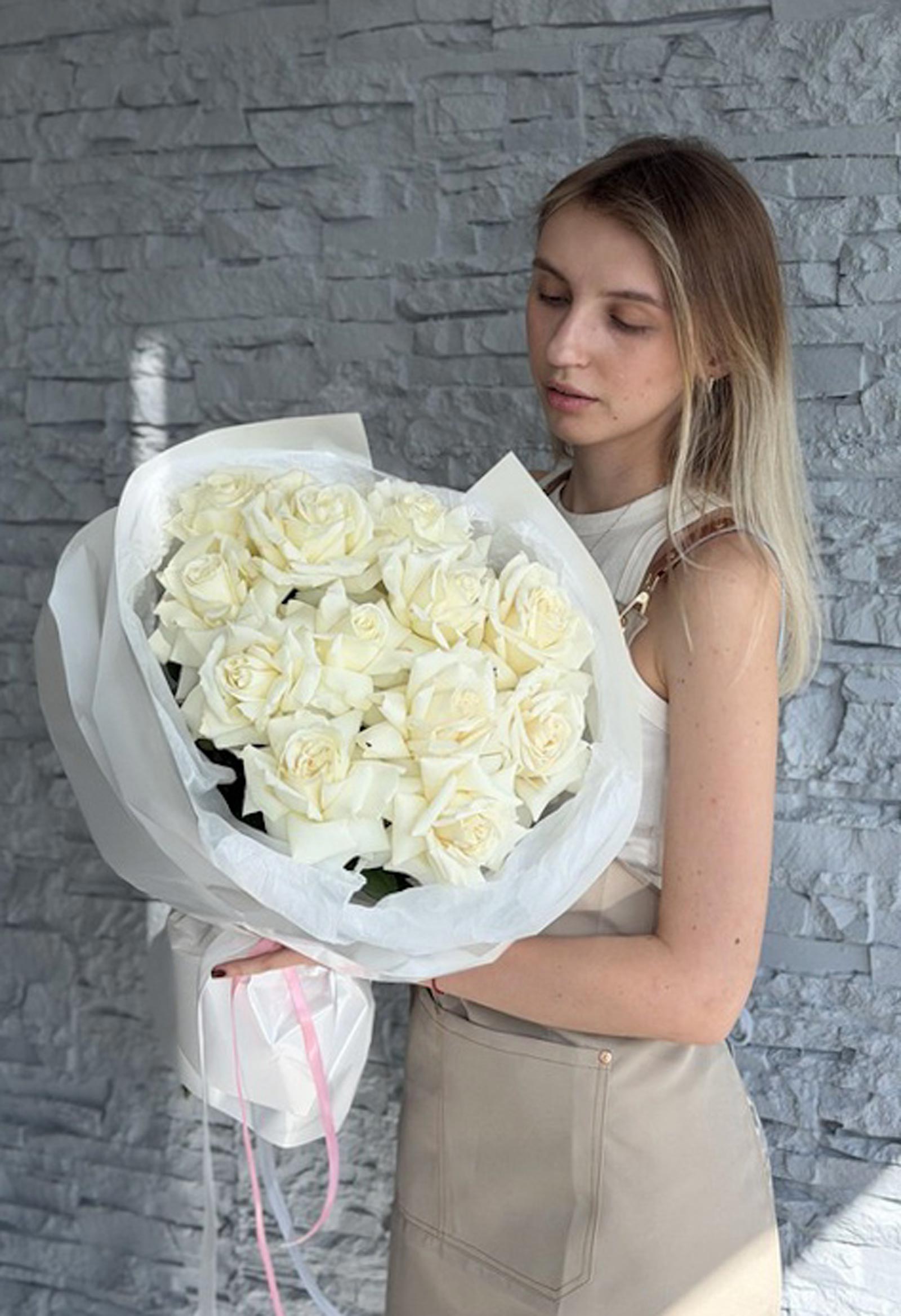 Woman holding a White French Roses bouquet wrapped in white paper against a stone wall.